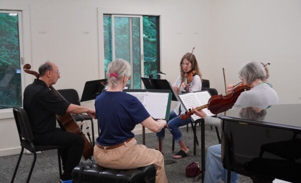 A small string ensemble rehearsing in a bright room, players reading sheet music with violins and a cello visible.