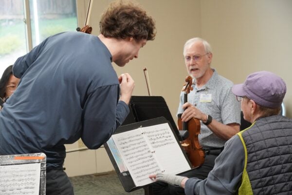 Musicians in a rehearsal room, a man with a violin standing over sheet music on a stand.